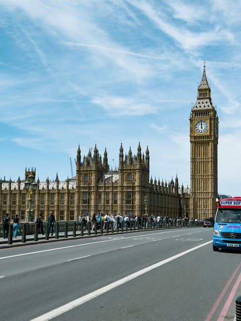 A wide view of a street in London showing the Houses of Parliament and the clock tower known as Big Ben under a partly cloudy sky. In the foreground, there is a paved road with white lane markings, a