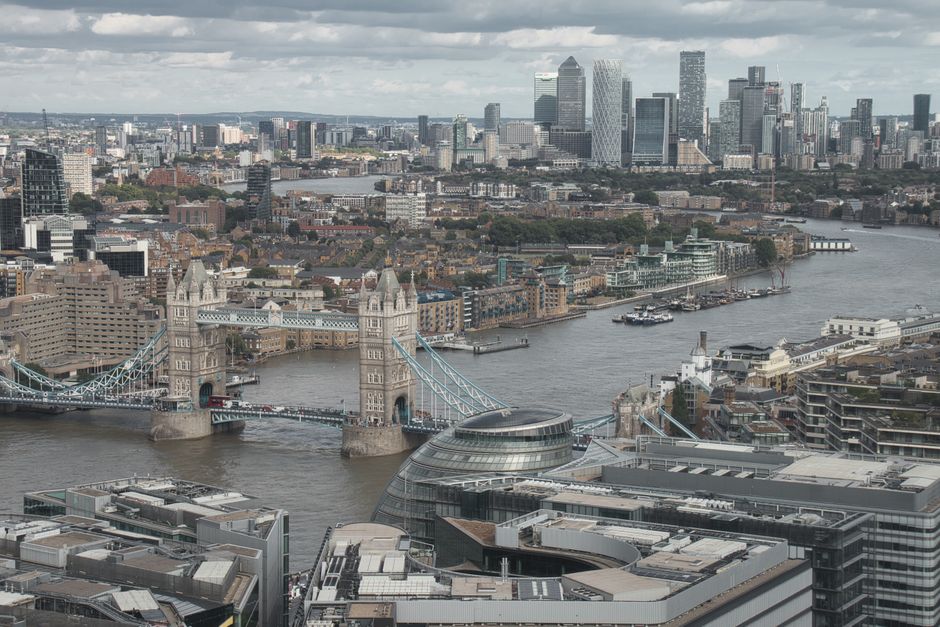 A wide view of a street in London showing the Houses of Parliament and the clock tower known as Big Ben under a partly cloudy sky. In the foreground, there is a paved road with white lane markings, a