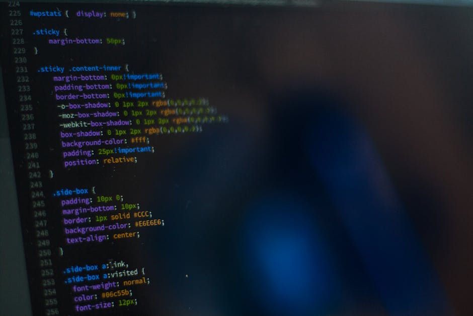 A computer monitor displays lines of programming code in various colours, including green, purple, and orange, on a dark background. The code appears to be related to software development, with visibl