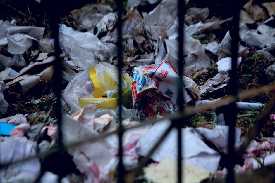 A close-up view of a pile of scattered household waste and packaging materials, including crumpled paper, plastic bags, and cardboard cartons, situated on the ground behind a black metal fence. The wa