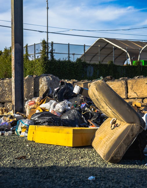 A close-up view of a pile of scattered household waste and packaging materials, including crumpled paper, plastic bags, and cardboard cartons, situated on the ground behind a black metal fence. The wa